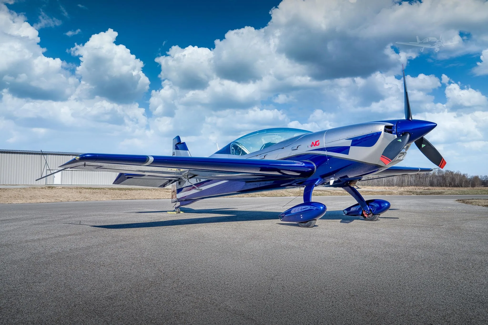 A blue and silver aerobatic aircraft, likely an Extra 330SC, is parked on an airfield tarmac under a partly cloudy sky.