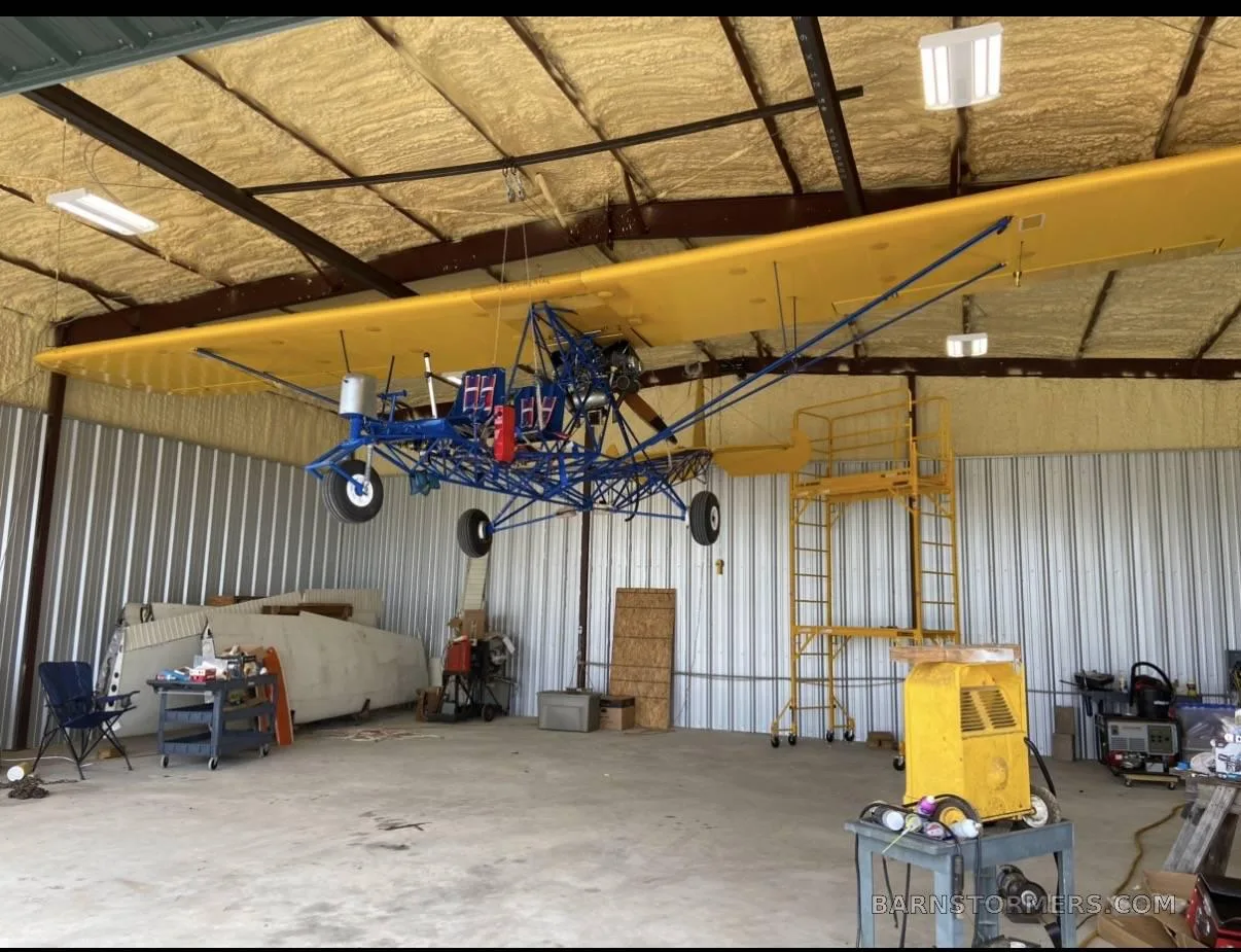 A yellow-winged, blue-framed ultralight aircraft is suspended from the ceiling inside a hangar.