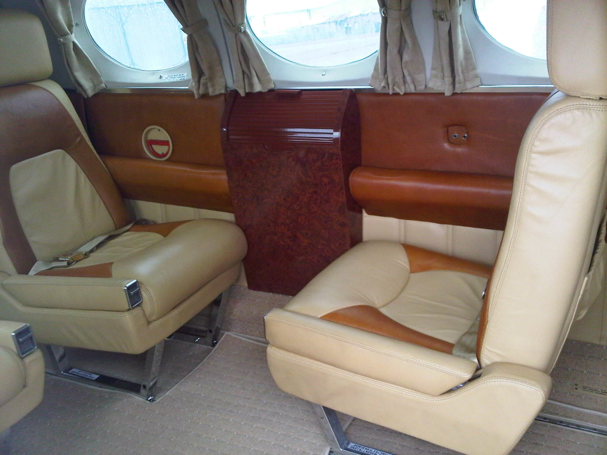 A view of the aircraft cabin showing two-tone tan and brown leather seats, a wood-paneled cabinet, and windows with curtains.