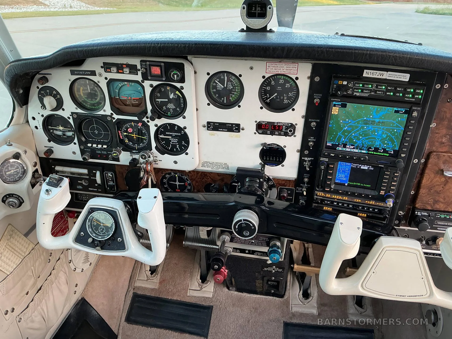 The cockpit of a Beechcraft Bonanza (N167JW) showing the instrument panel with analog gauges, a modern GPS display, and white control yokes.