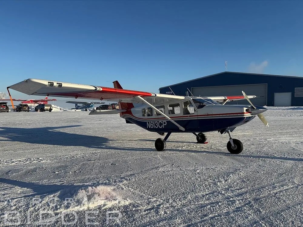 A blue and white Cessna 206 with red accents, registration N613CP, parked on a snowy tarmac in front of a hangar.