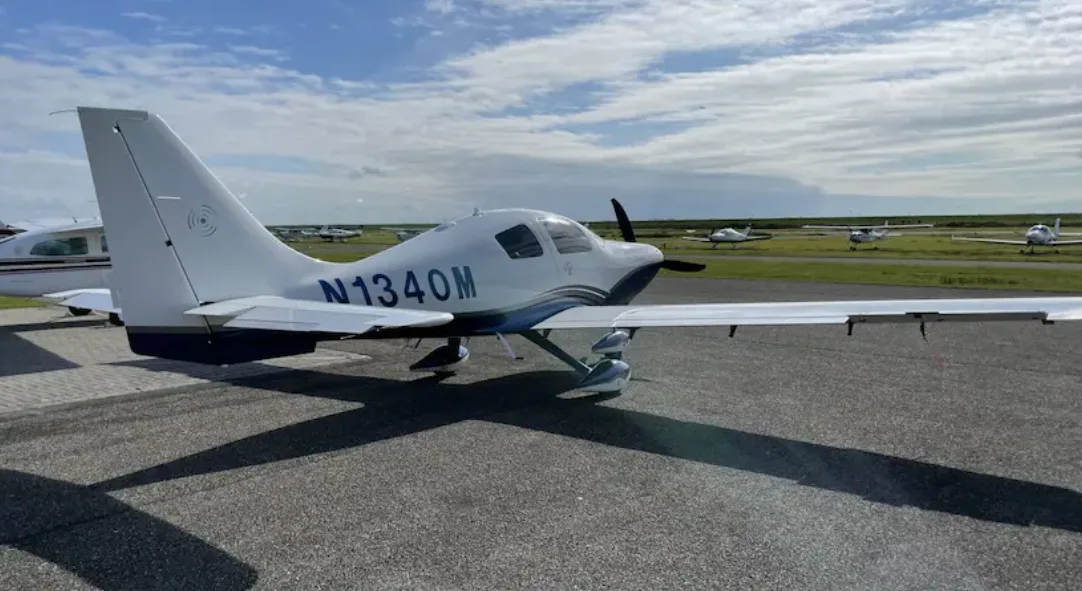 A white and blue Columbia 400 aircraft with registration N1340M parked on an airport tarmac with other planes in the distance.