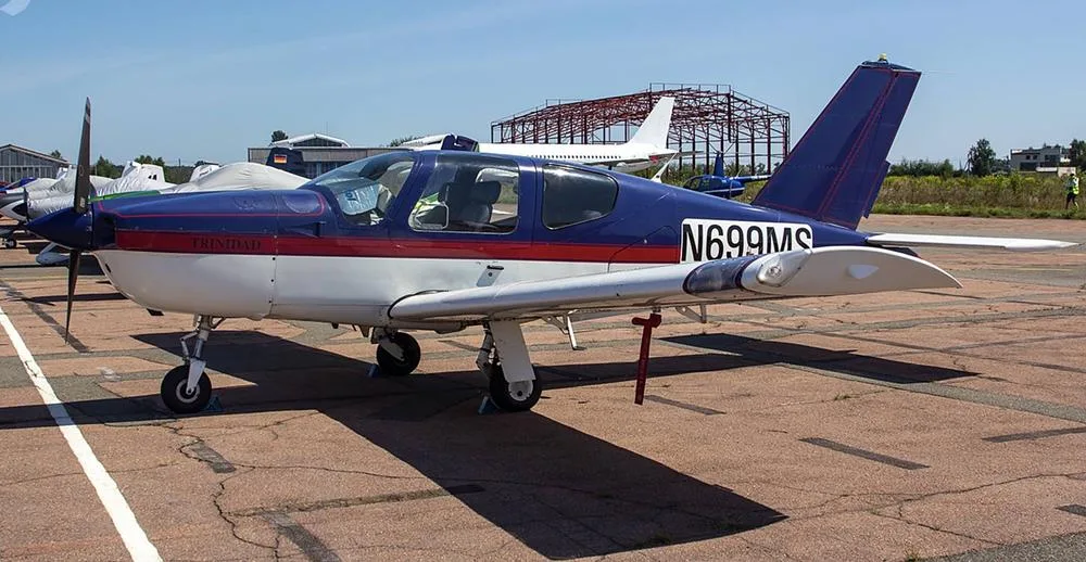 A blue and white Socata Trinidad aircraft, N699MS, is parked on an asphalt ramp with a propeller cover.