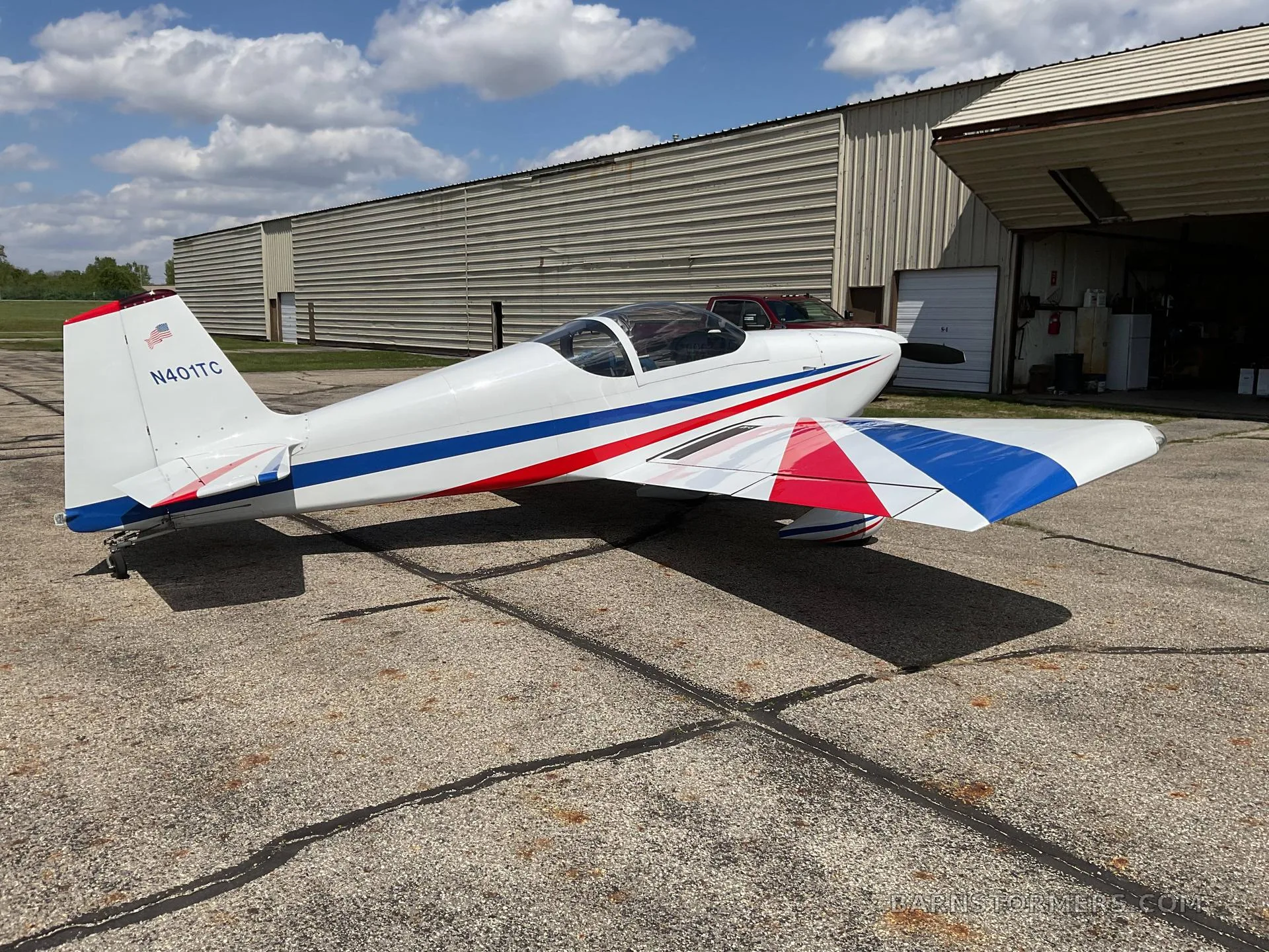 A white single-engine aircraft with red and blue stripes on its wings and tail (N401TC), parked on an asphalt tarmac with a hangar in the background.