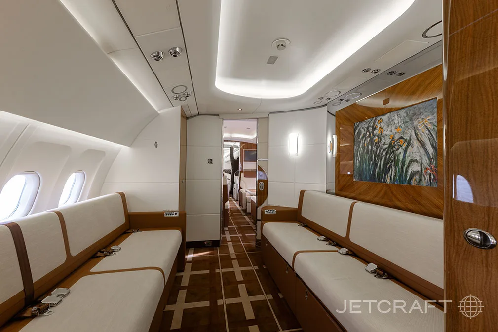 A view down the aircraft cabin aisle, showing two long sofas and wood paneling with a large artwork.