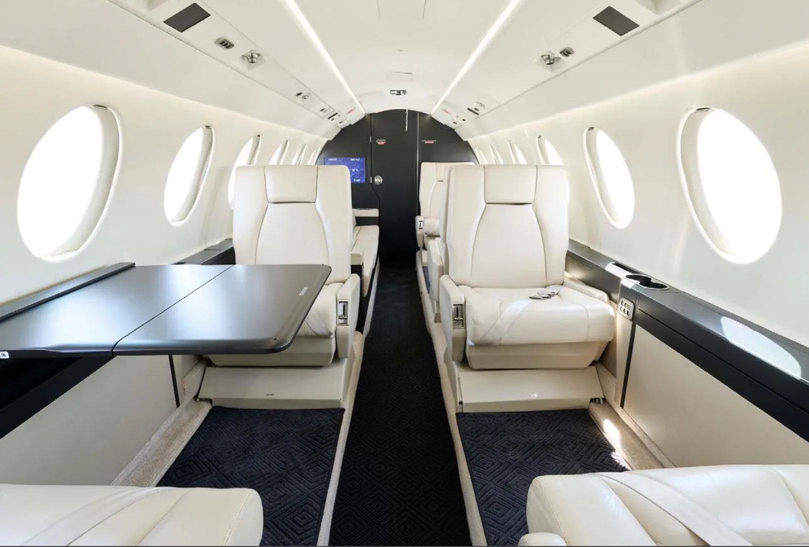 The luxurious interior of a private jet cabin features light-colored leather seats, a fold-out table, and large oval windows.