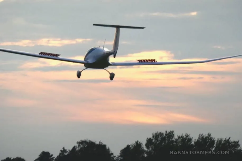 A white motor glider with airbrakes extended is silhouetted against a sunset sky over trees.