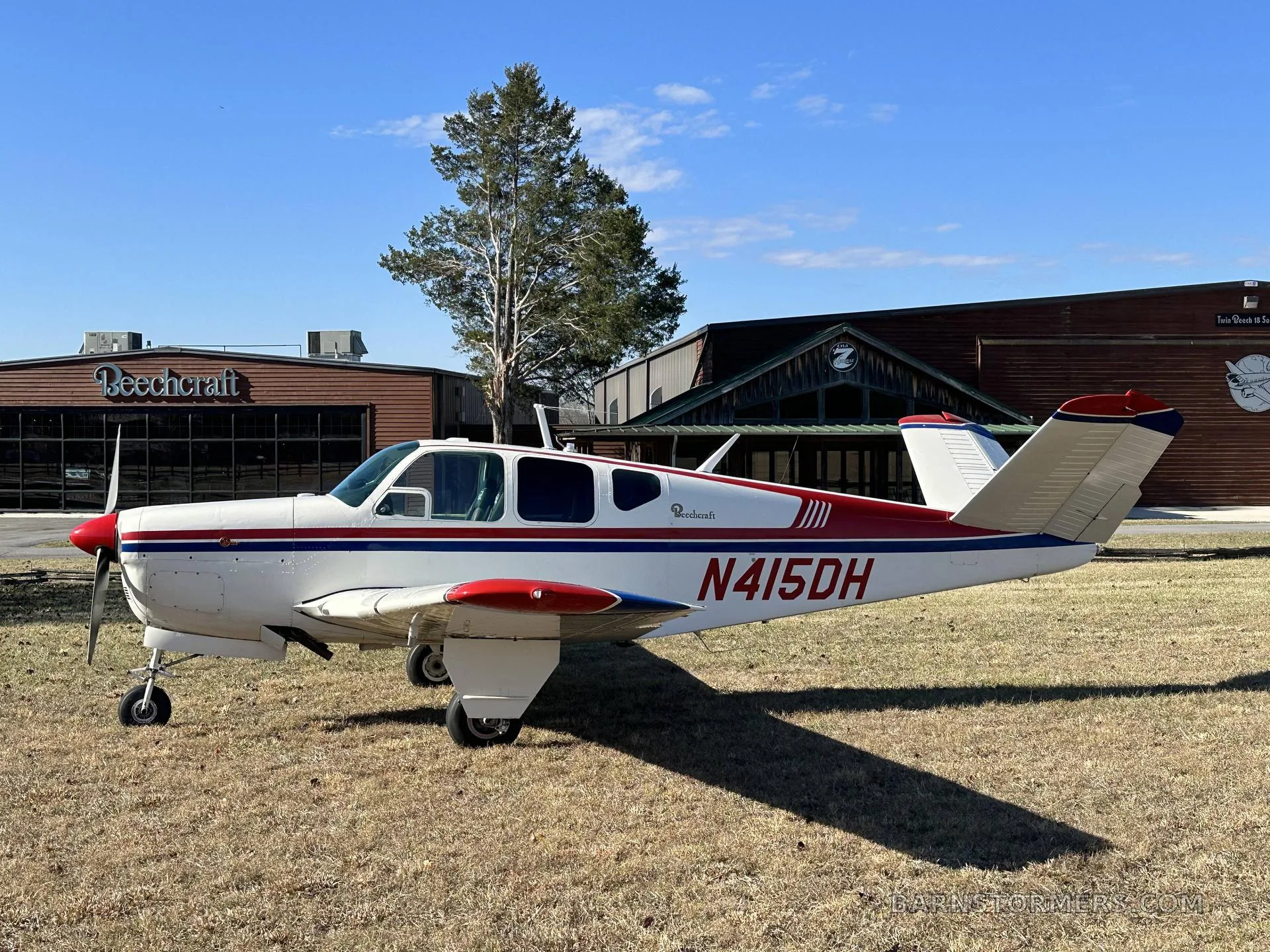 A white and red Beechcraft Bonanza N415DH parked on grass in front of a building with a Beechcraft sign.