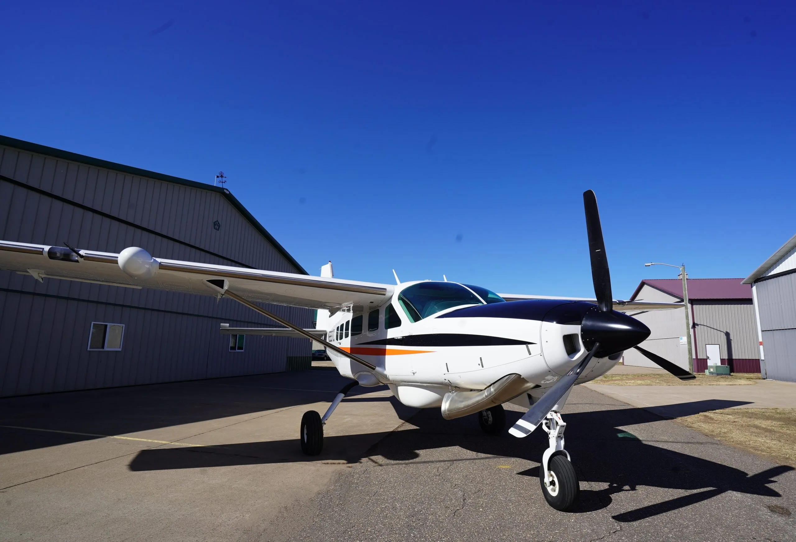 A white Cessna Caravan with black and orange stripes parked on an airfield tarmac next to hangars under a clear blue sky.