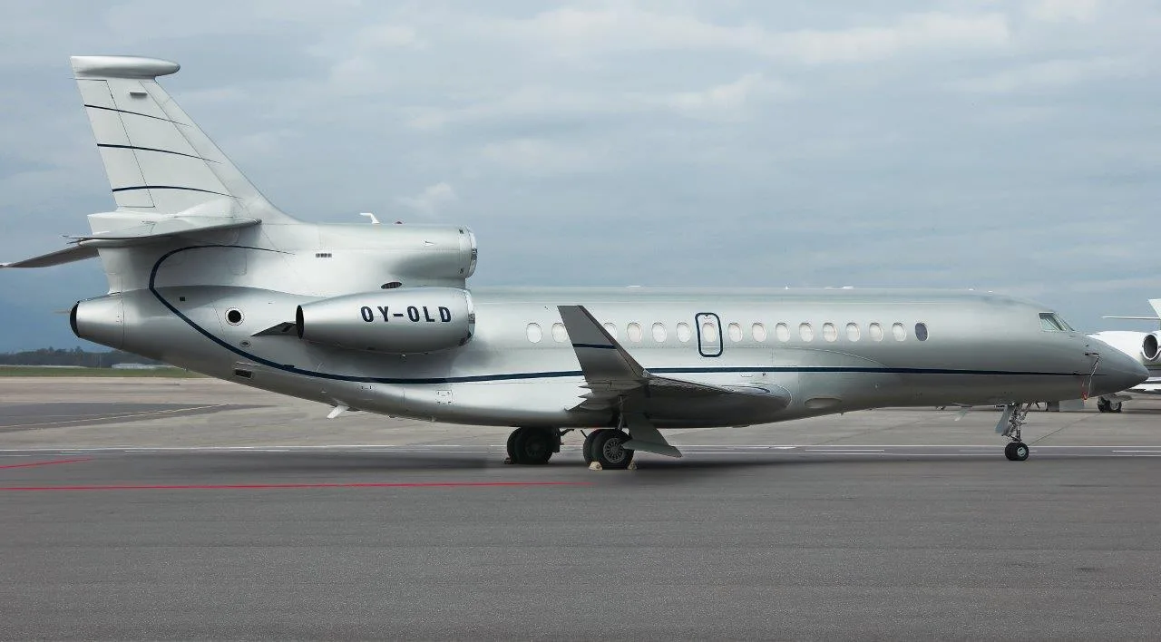 A silver Dassault Falcon business jet with registration OY-OLD is parked on an airport tarmac under a cloudy sky.