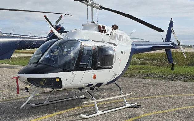 A white and blue Bell 206 JetRanger helicopter is parked on an asphalt tarmac with another helicopter in the background.