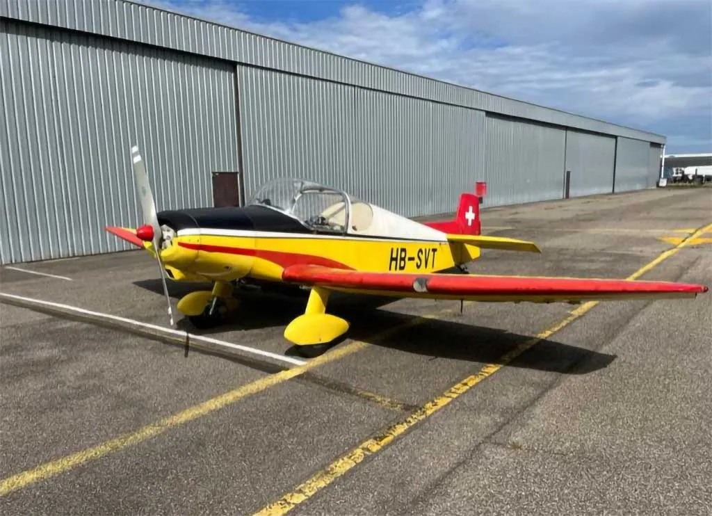 A yellow, red, and black single-engine aircraft (HB-SVT) parked on an asphalt tarmac in front of a metal hangar.
