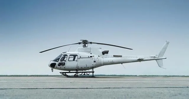 A white single-engine helicopter with skid landing gear sits on an asphalt runway under a clear sky.