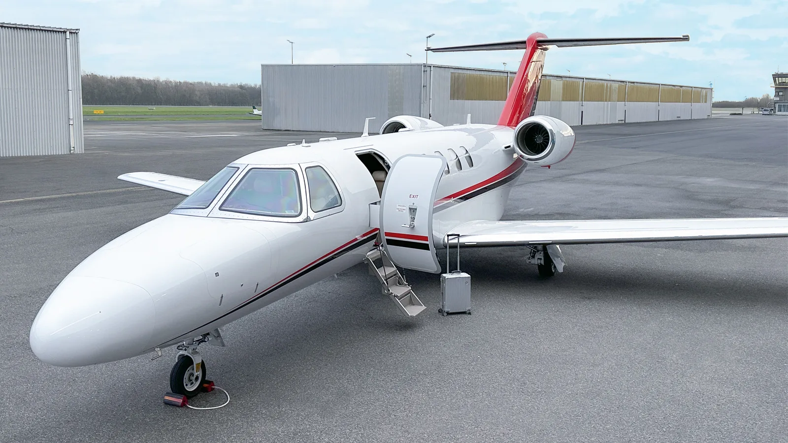 A white Citation CJ4 business jet with red and black stripes on the tarmac, seen from a slightly higher angle, with its main cabin door open and a suitcase nearby.