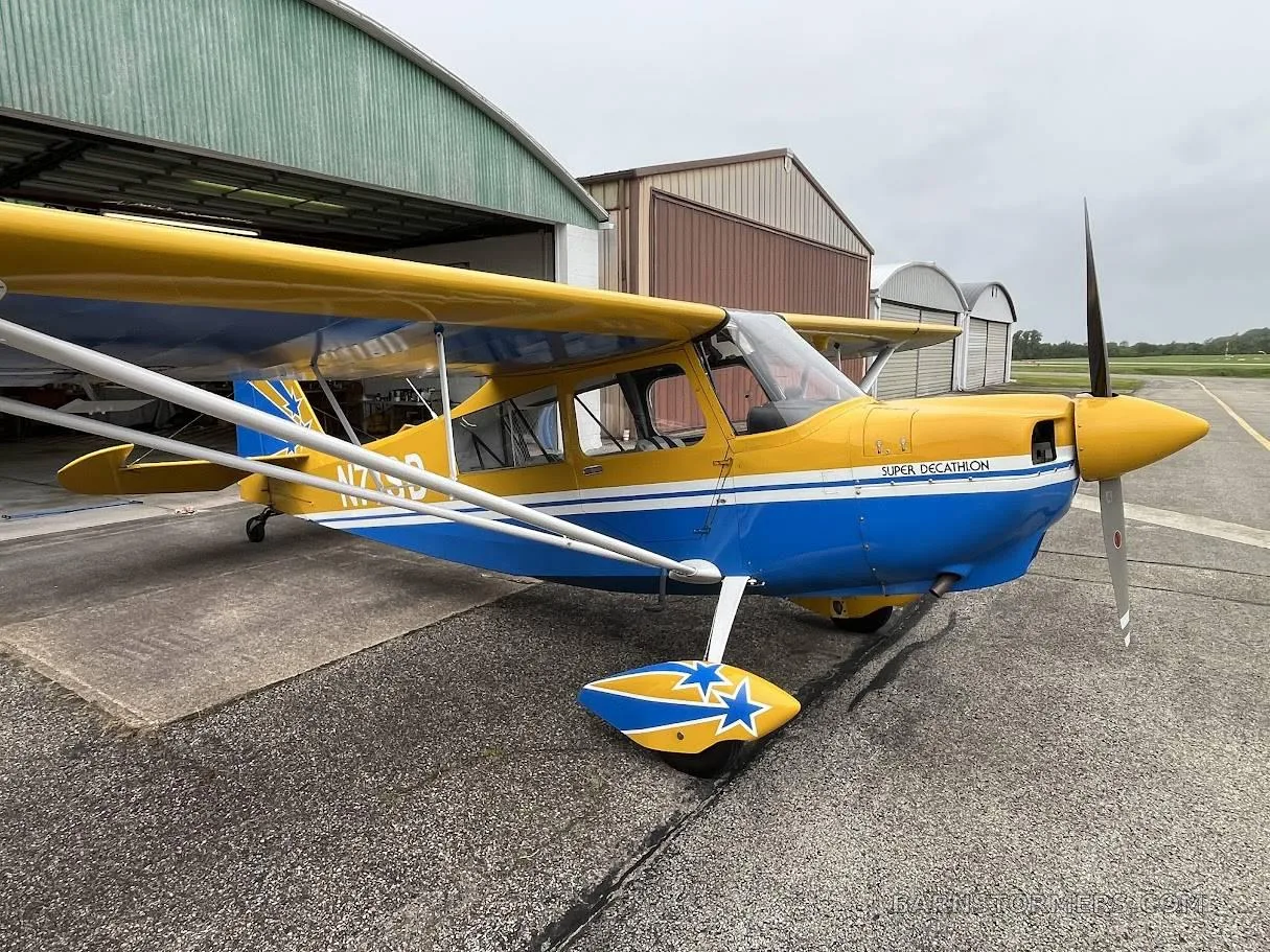 A yellow and blue Super Decathlon aircraft with tail number N71SD parked in front of an open hangar, viewed from the front-right.