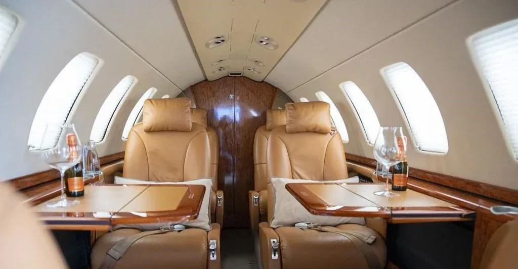 The luxurious interior cabin of a private jet with tan leather seats, wooden tables, and champagne glasses.