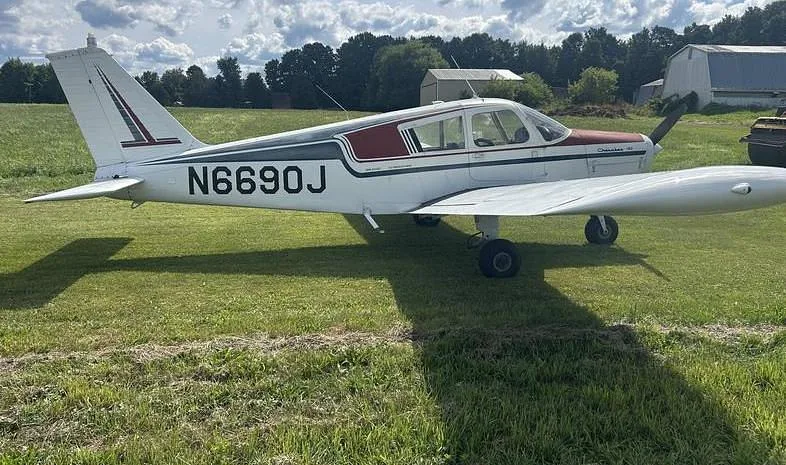 A white and maroon Piper Cherokee 140 aircraft, N6690J, parked on a grassy field under a partly cloudy sky.