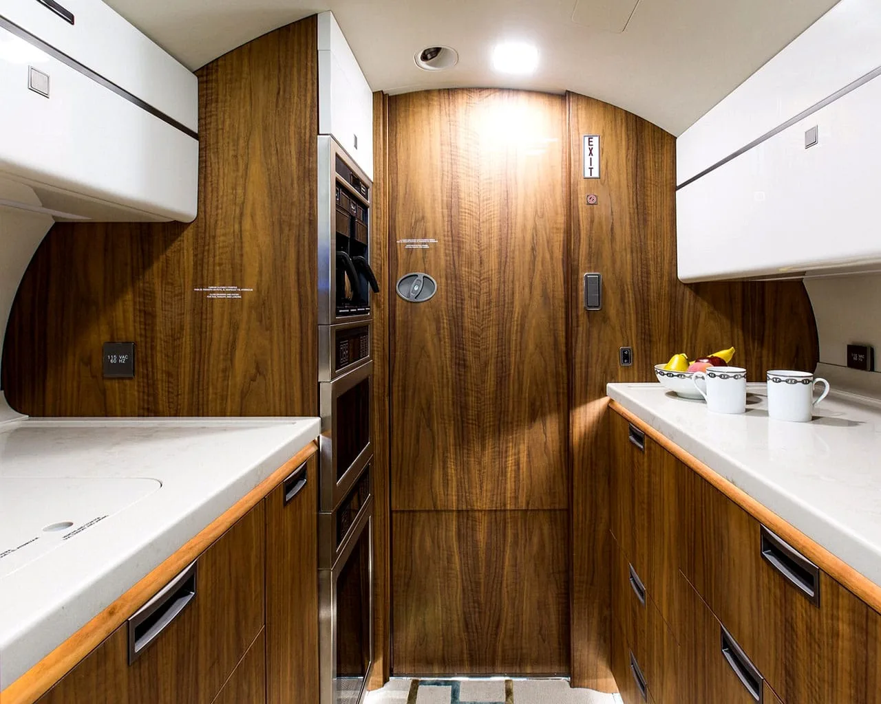 The galley area of a private jet, featuring white countertops, dark wood cabinetry, and built-in appliances.