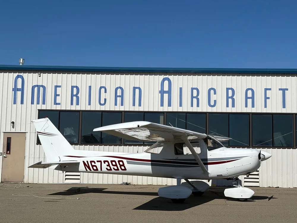 A white Cessna 172 with red and gray stripes, registration N67398, parked on an airfield in front of an "American Aircraft" hangar.