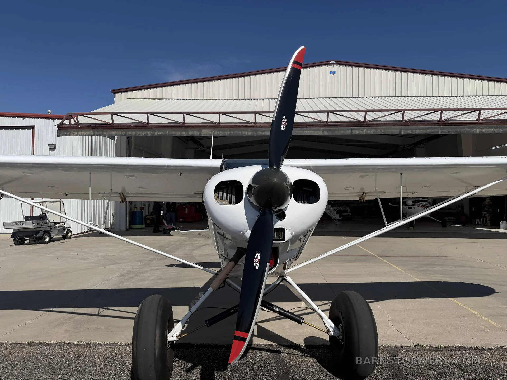 A head-on view of a white single-engine bush plane with a black propeller and large tires, parked in front of a hangar.