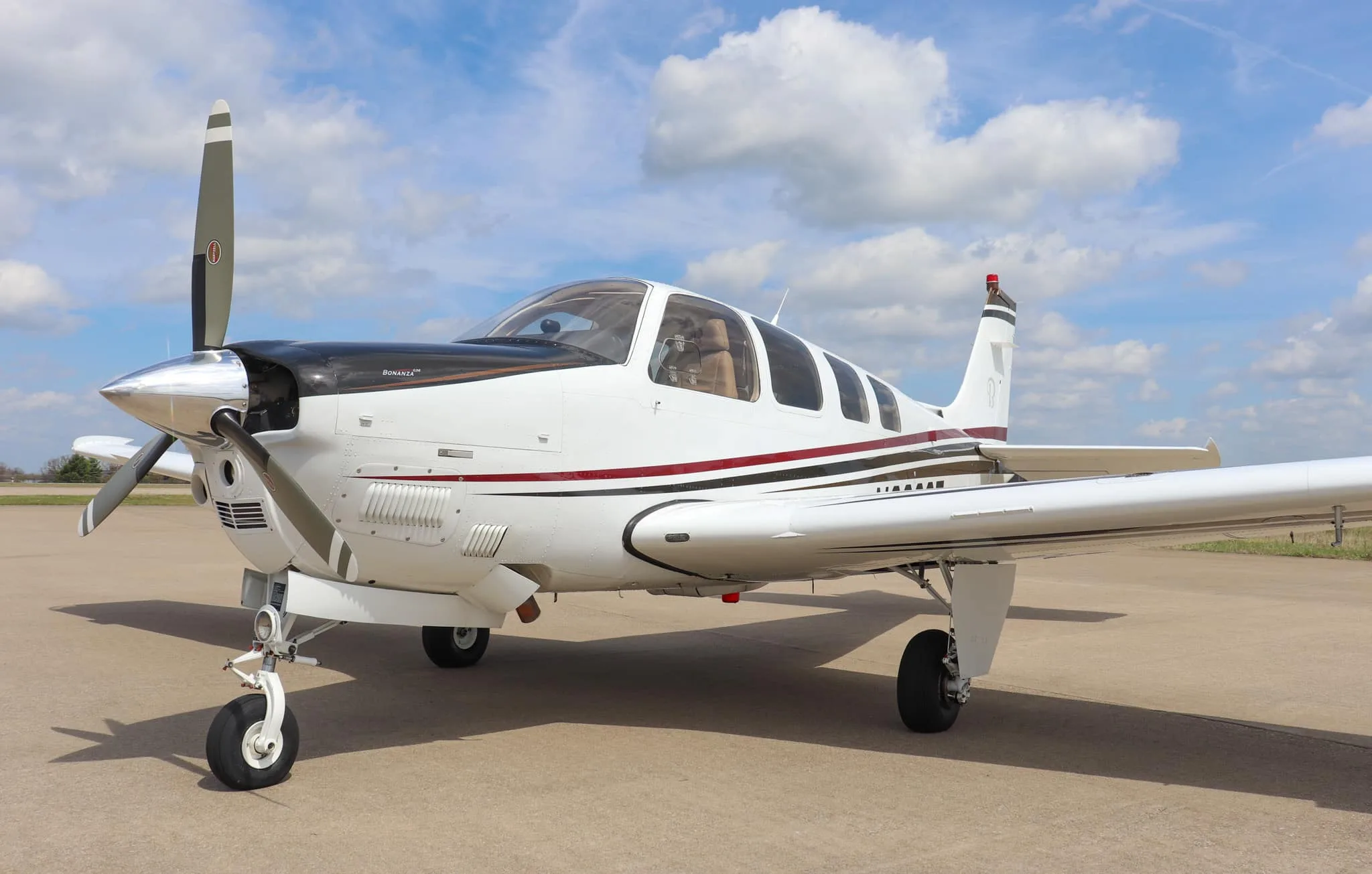 A front-quarter view of a white Beechcraft Bonanza G36 aircraft with black, red, and brown stripes, parked on an airfield.