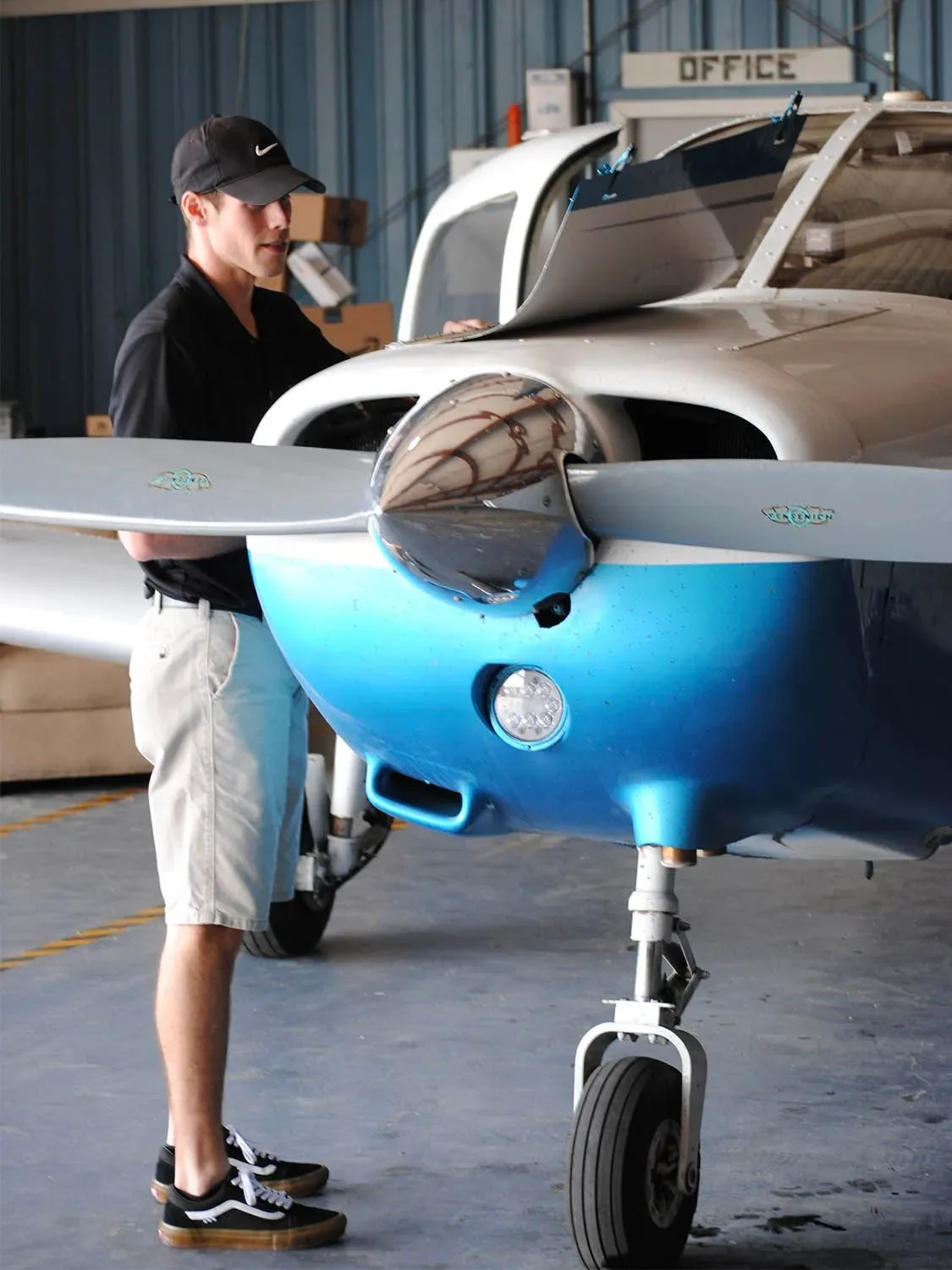 A person in a black cap and shirt inspecting the engine of a blue and white single-engine aircraft in a hangar.