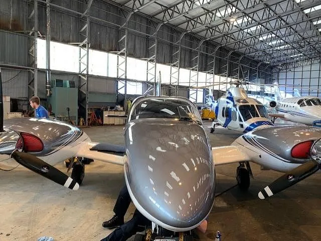 Front view of a grey and white twin-engine aircraft with blue stripes inside a hangar, with other aircraft in the background.