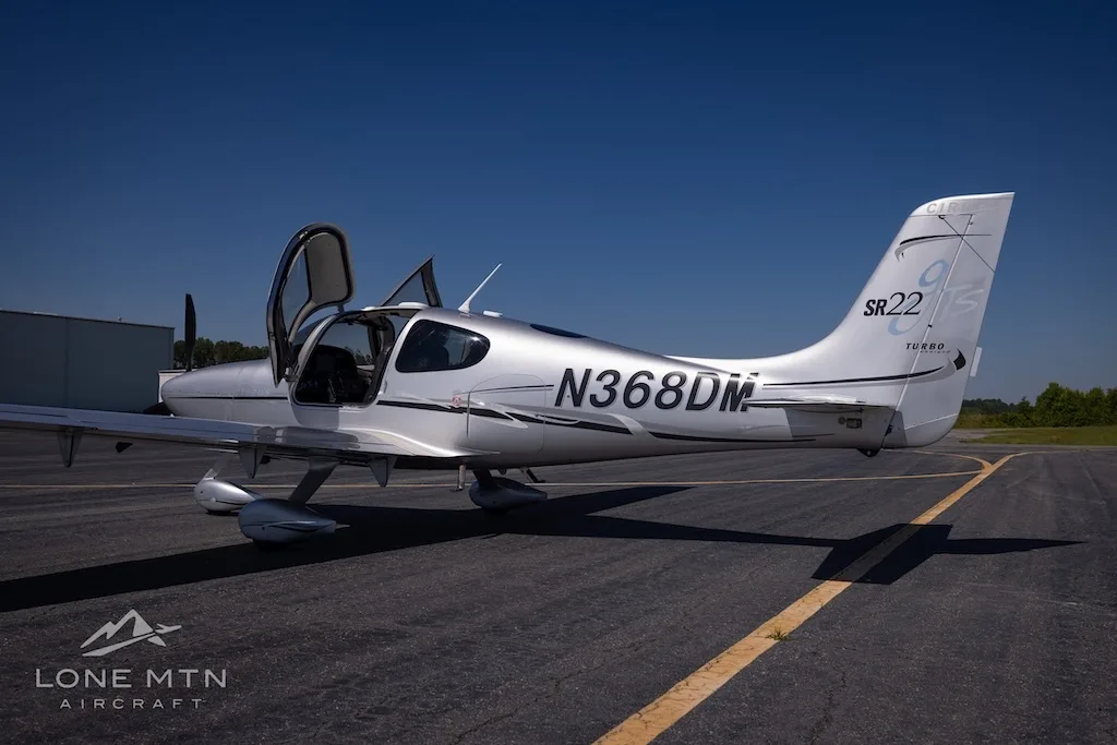 A silver Cirrus SR22 Turbo aircraft (N368DM) with its doors open, parked on an asphalt runway under a clear blue sky.