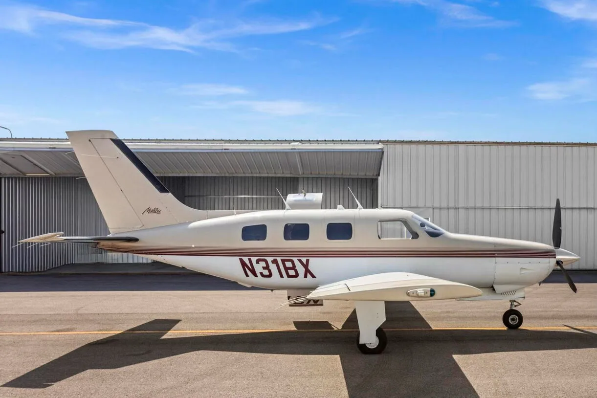A full side view of the Piper Malibu N31BX, showing its white and tan paint scheme with red stripes, parked on an airfield.