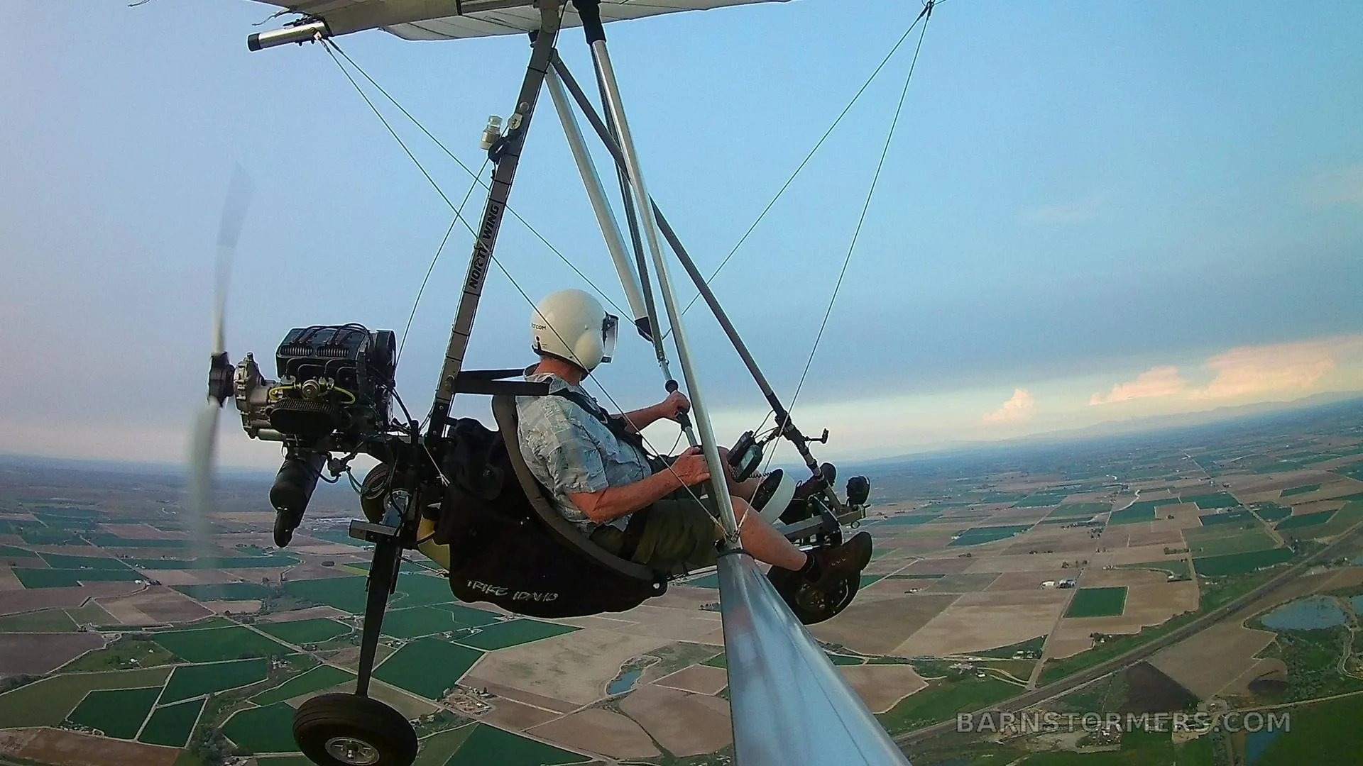 A pilot in a white helmet flies a black and white ultralight aircraft over green and brown fields under a blue sky.