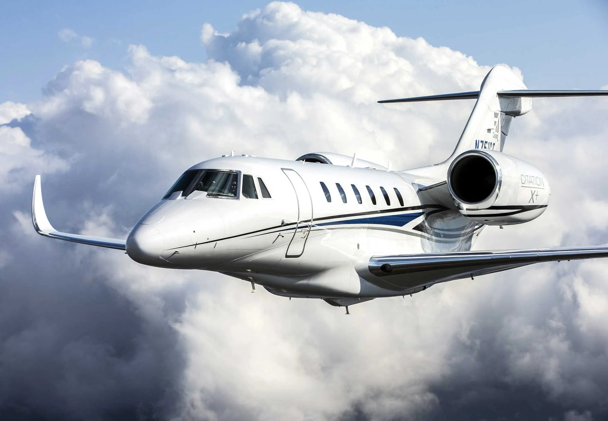 A white Cessna Citation X+ business jet with blue and black stripes flies through a cloudy sky.
