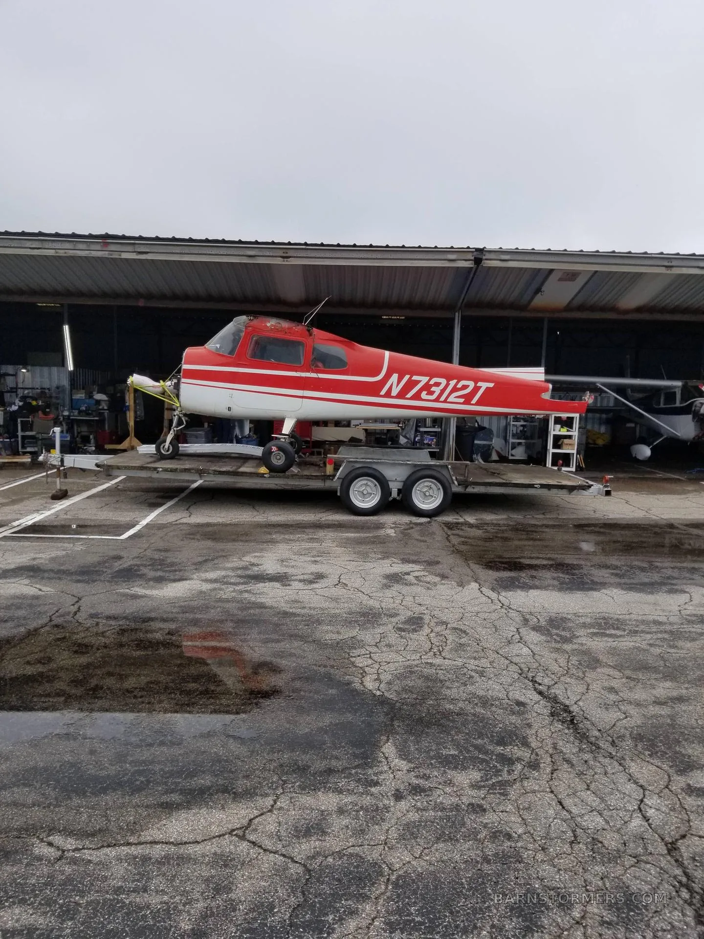 A red and white single-engine aircraft, N7312T, with its engine cowling removed, sits on a trailer outside a hangar on a wet tarmac.