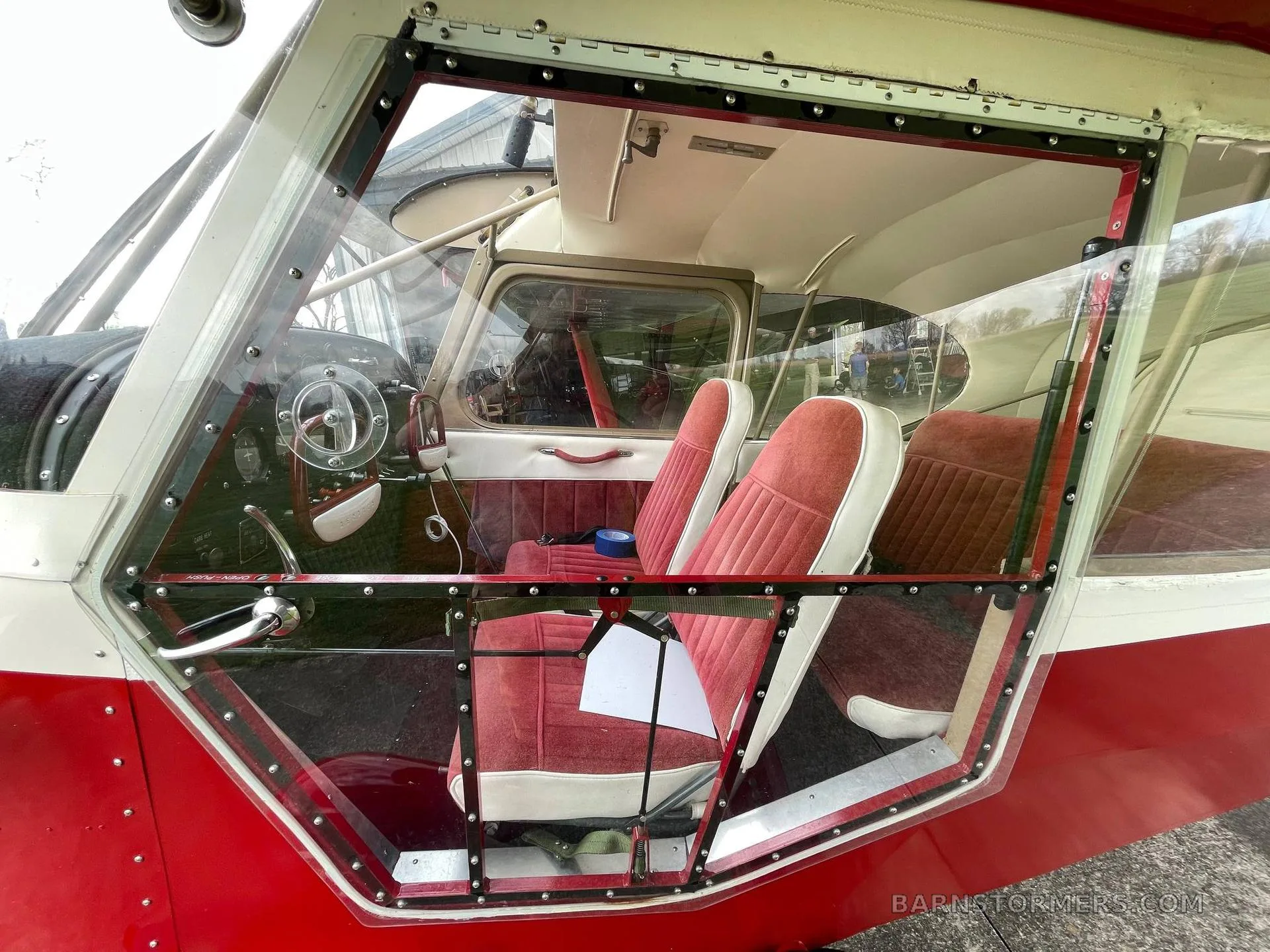 The red and white interior of a vintage aircraft, showing two front seats and a view of the cockpit through the side window.