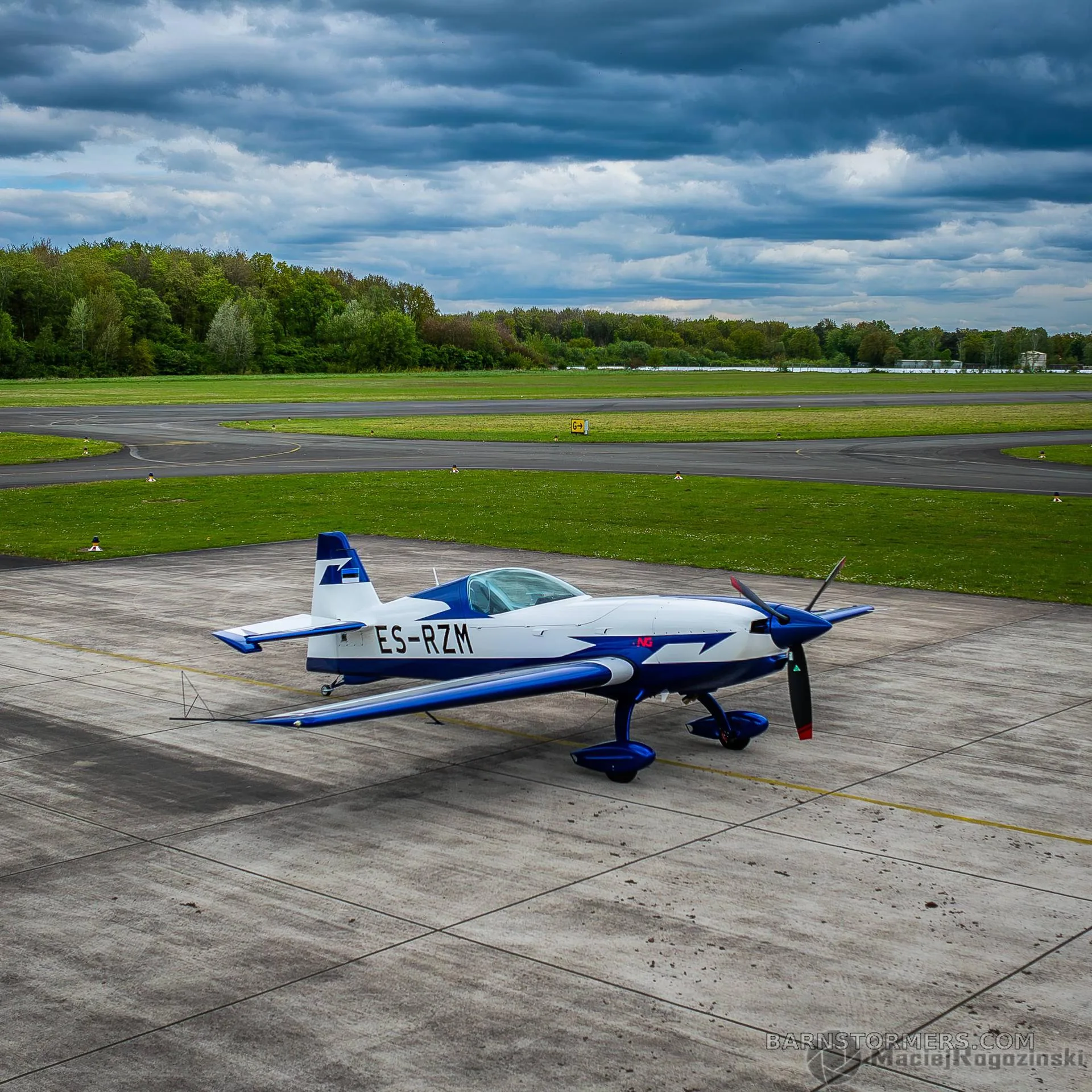 A white and blue Extra 330SC aircraft with registration ES-RZM is parked on an airfield under a cloudy sky.