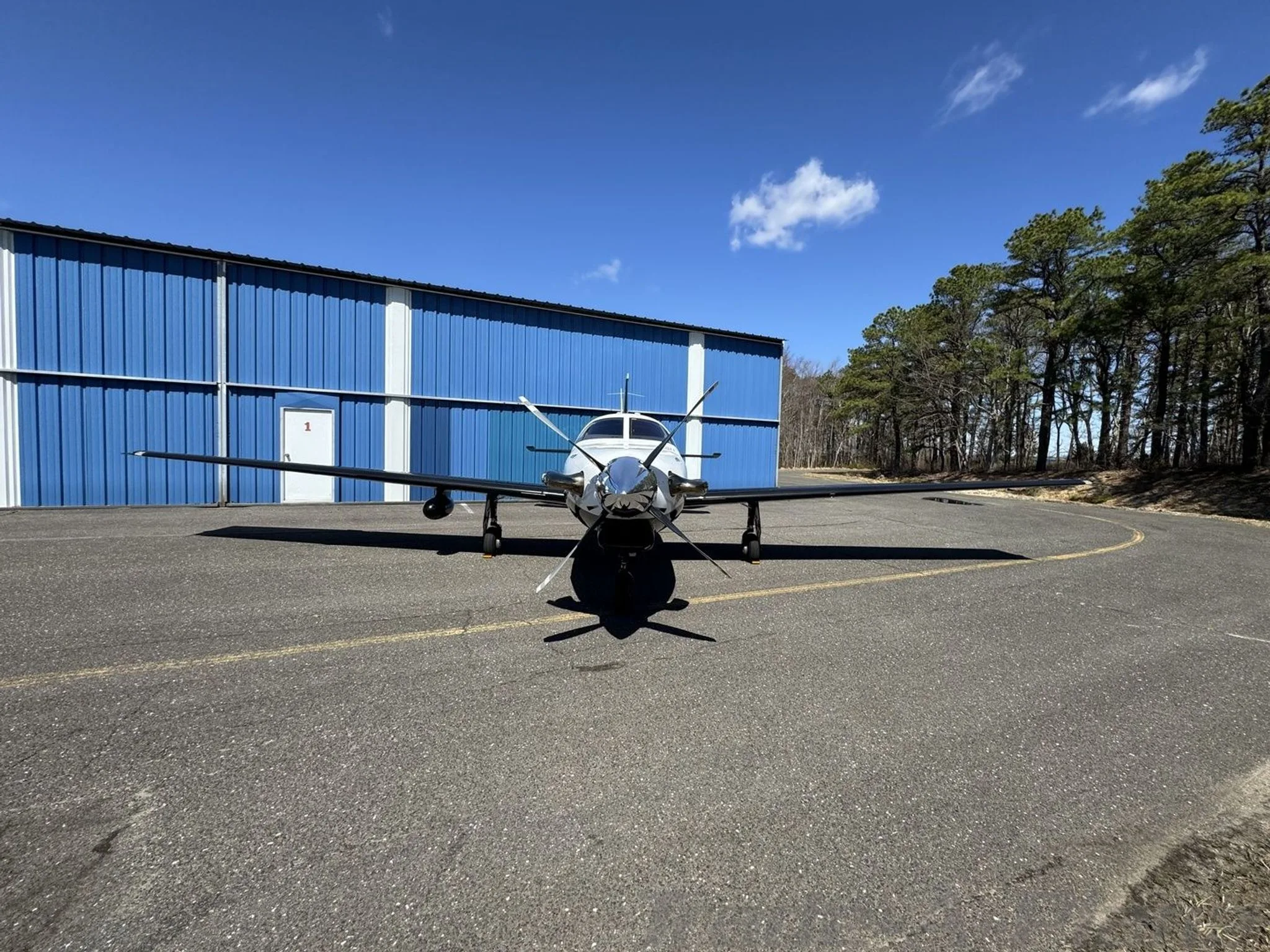 A front view of a white and black Piper M-class aircraft parked on an asphalt ramp in front of a blue hangar.