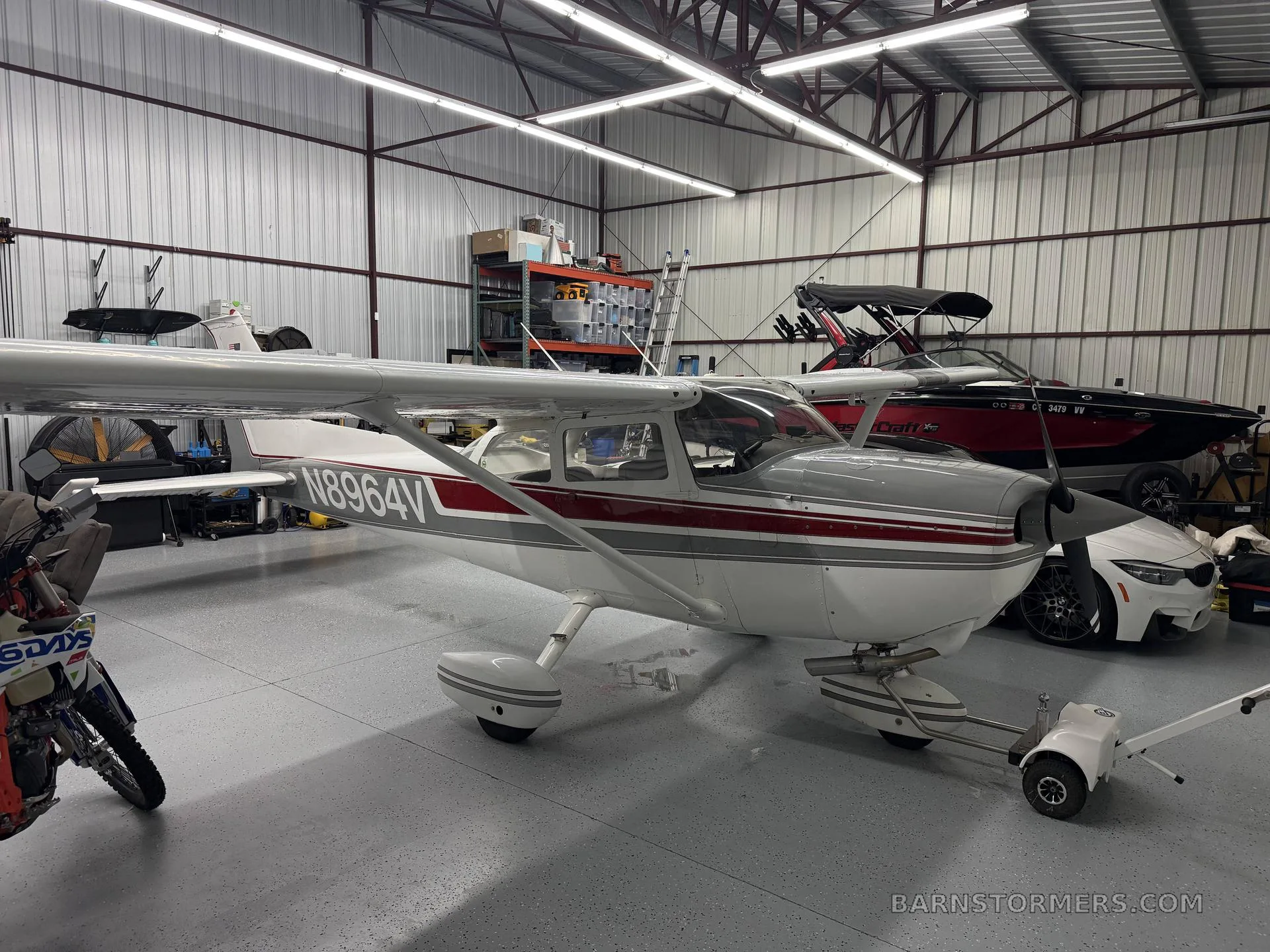 A white and grey Cessna 172M with red stripes, registration N8964V, parked in a hangar next to a red boat and a white car.