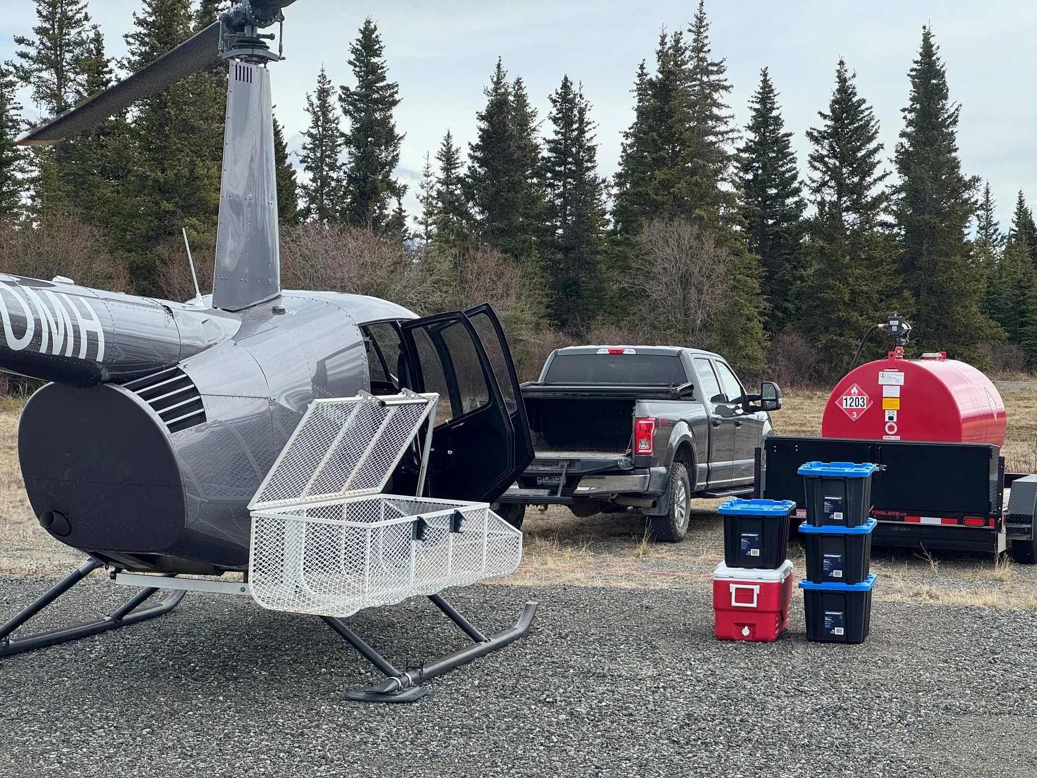 A grey Robinson R44 Raven II helicopter with an open door and cargo basket, parked on gravel next to a pickup truck and a red fuel tank.