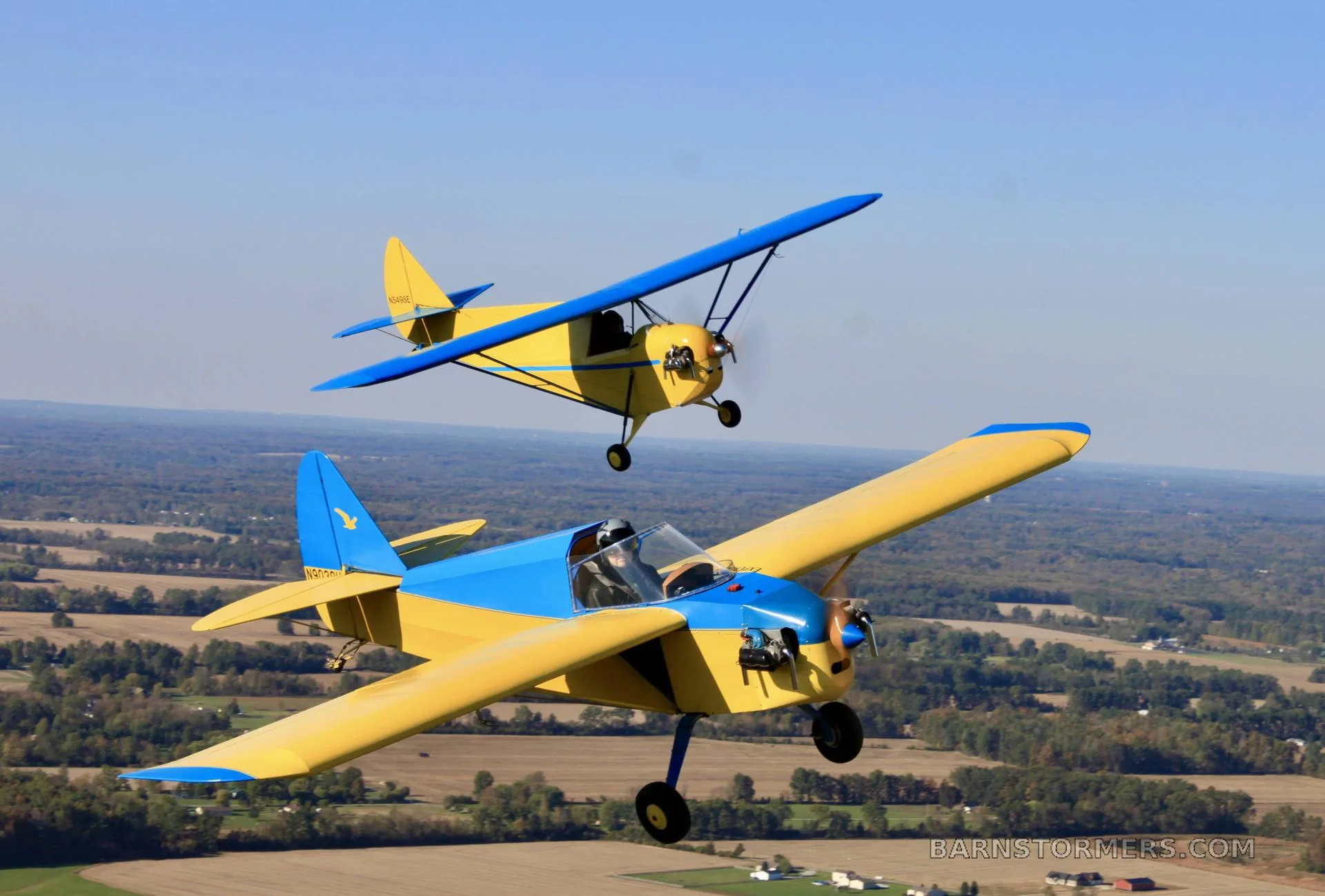 Two yellow and blue single-engine aircraft flying in formation over a rural landscape.