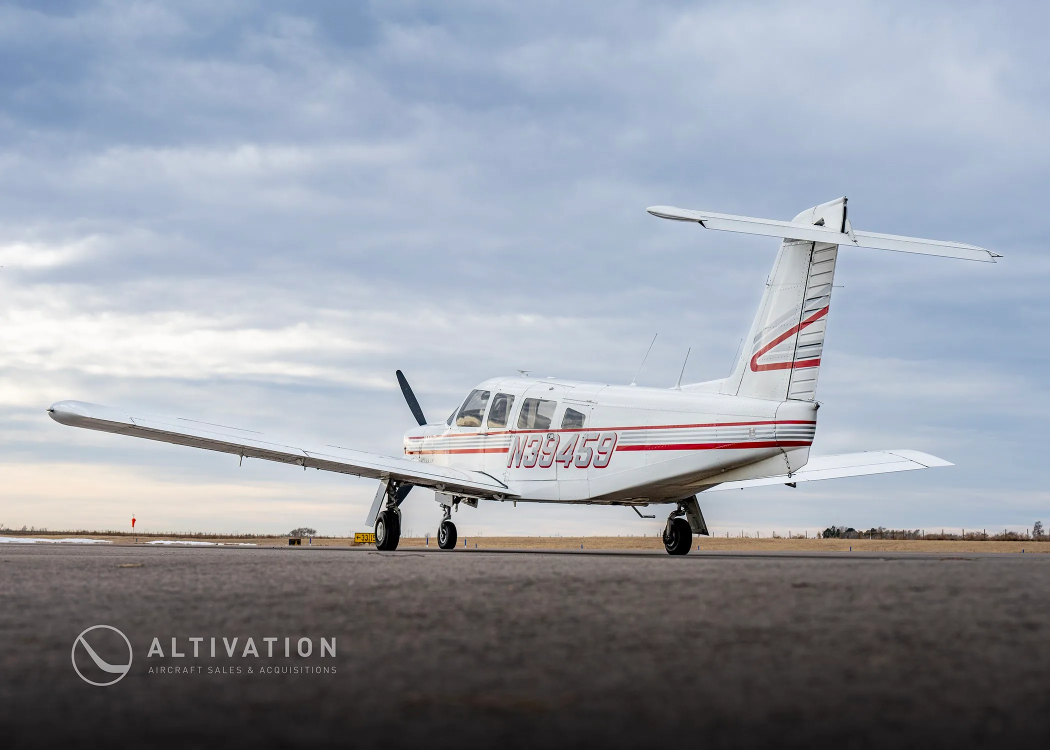 A white single-engine propeller aircraft with red and grey stripes, tail number N39459, seen from the rear-side on an airfield.