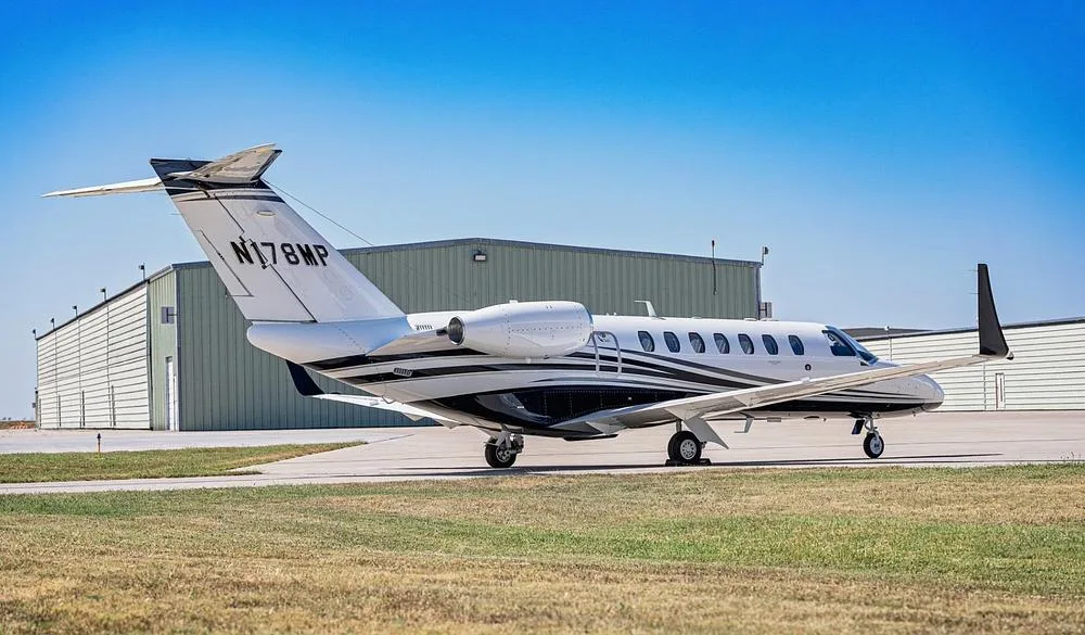 A side view of the white and blue Cessna Citation M2 jet, N178MP, parked on an airport tarmac with hangars in the background.