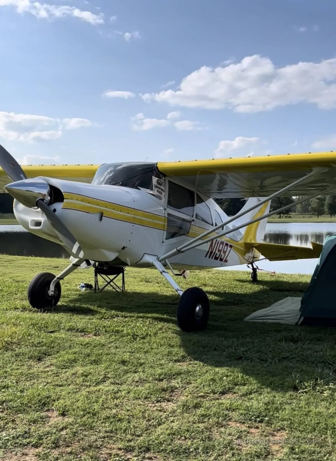 A white and yellow Maule aircraft (N199Z) is parked on a grassy field beside a lake under a partly cloudy sky.
