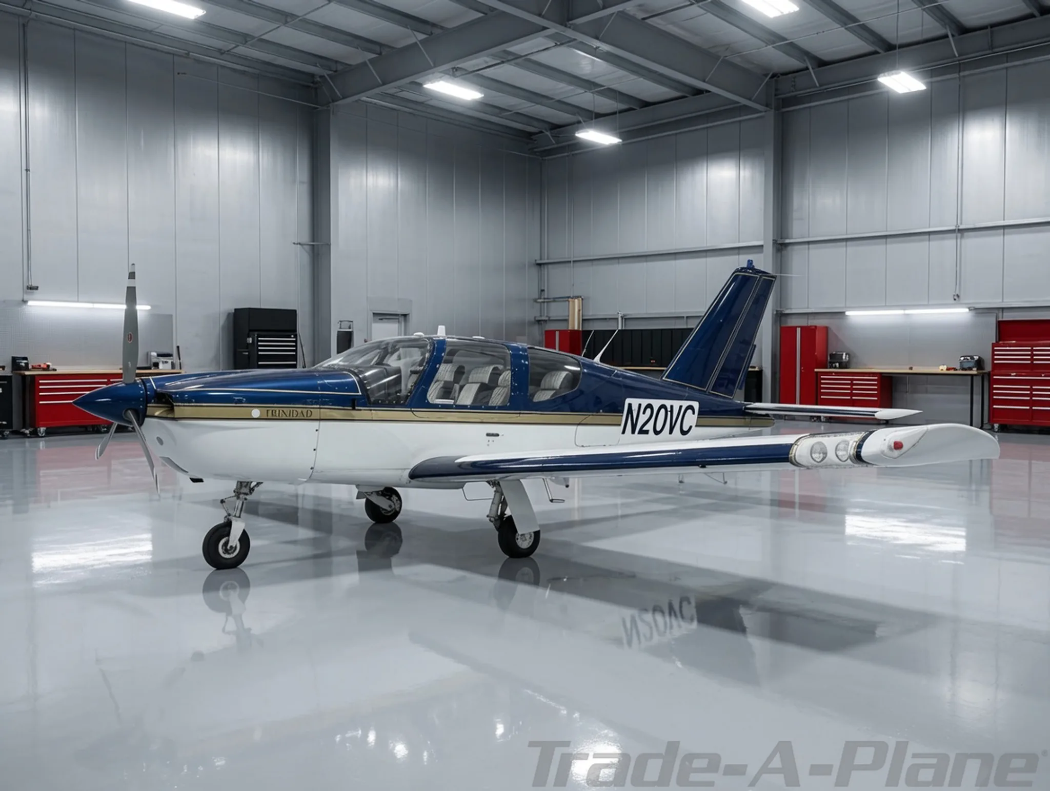 A white and blue Socata Trinidad TB20 aircraft with gold trim, tail number N20VC, parked inside a modern hangar.