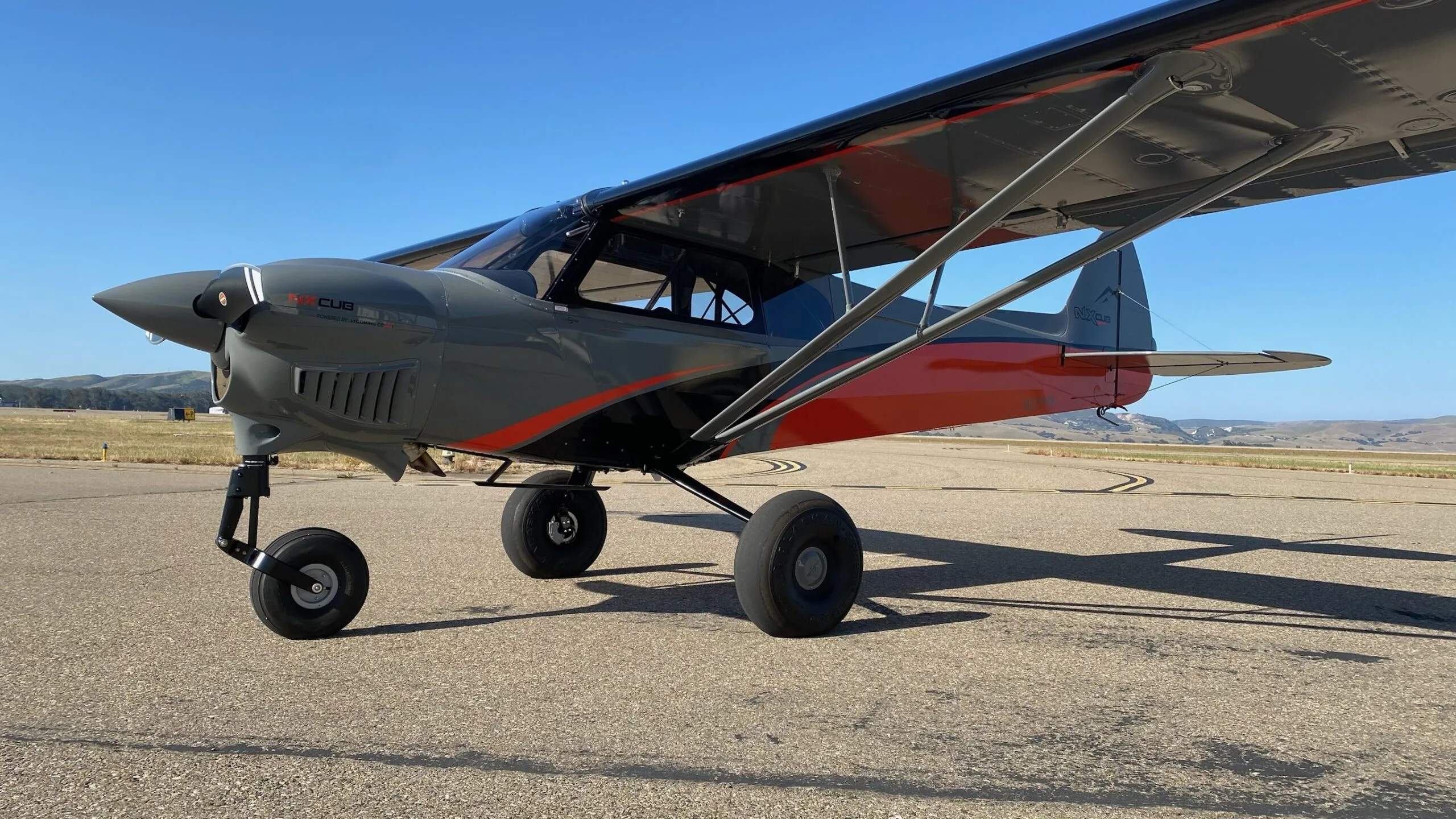 A gray and orange NX Cub aircraft with large tundra tires is parked on an airfield under a clear blue sky.