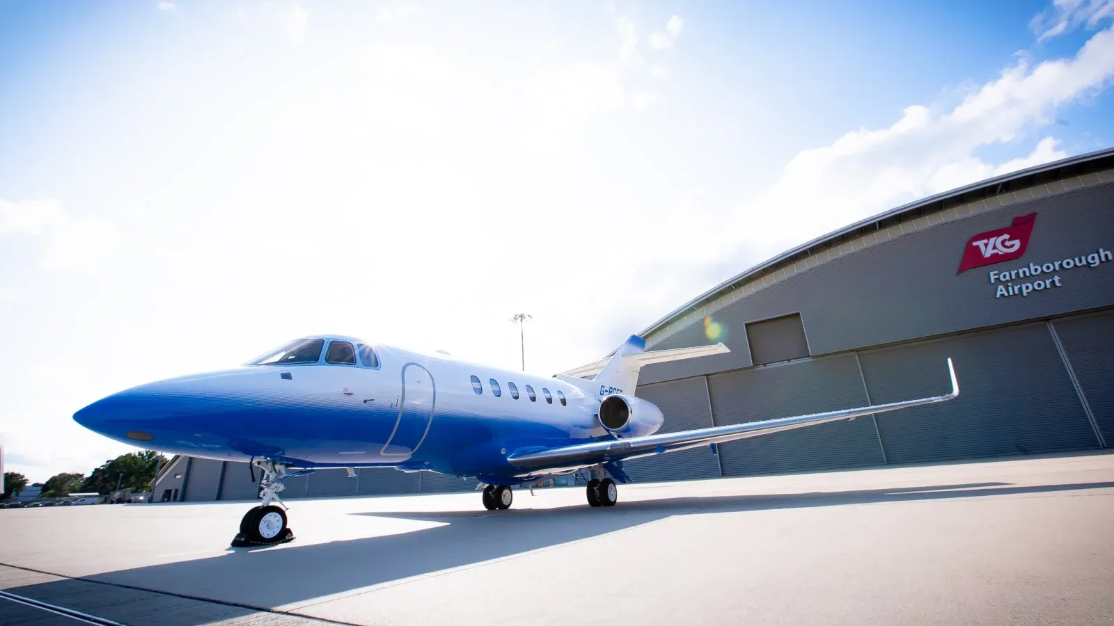 A blue and white jet sits on the tarmac in front of a hangar at Farnborough Airport.