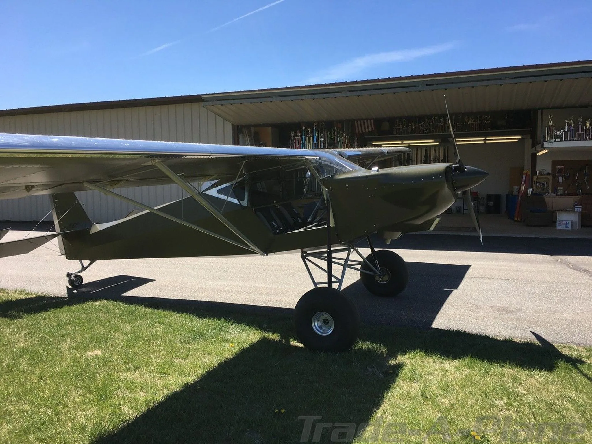 Side view of a green experimental aircraft with large tires parked outside a hangar on a sunny day.