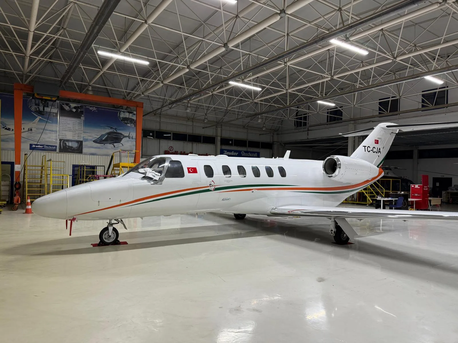 A white business jet with orange and green stripes and Turkish flag tail number TC-CJA, parked in a hangar.