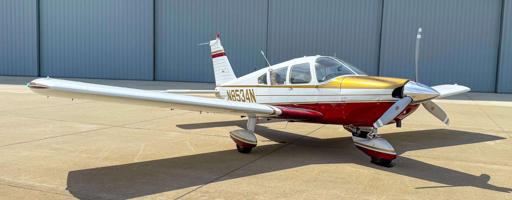 A white, red, and gold Piper Cherokee aircraft (N8534N) parked on an airfield in front of a hangar.