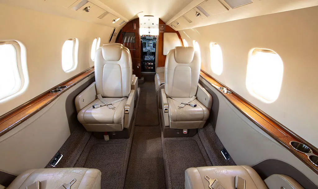 The luxurious interior cabin of a private jet with light beige leather seats and wood trim, looking towards the cockpit.