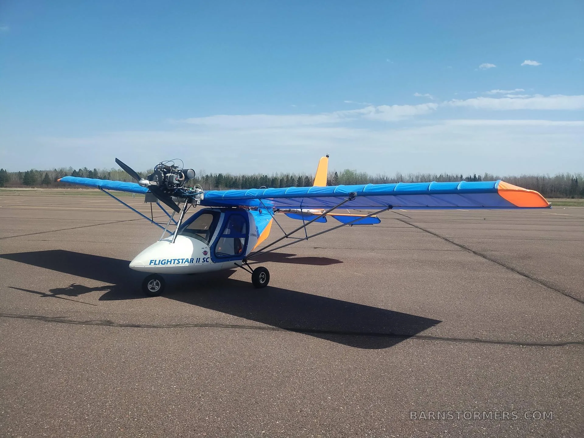 A blue and orange Flightstar II SC ultralight aircraft parked on an asphalt runway under a blue sky.
