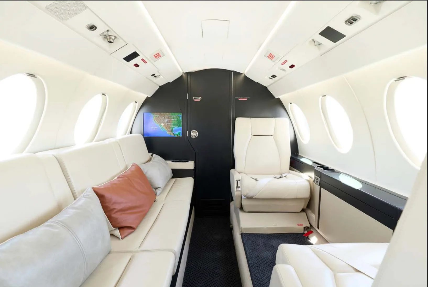 A view inside a private jet cabin shows a comfortable cream-colored sofa, individual seats, and a small screen displaying a map.